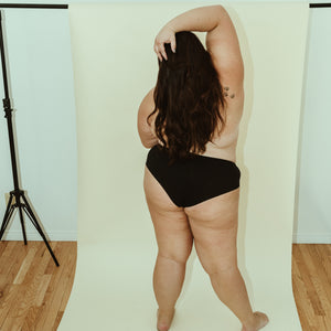 Plus size woman posing in onyx (black) cheeky organic cotton underwear, rear view.